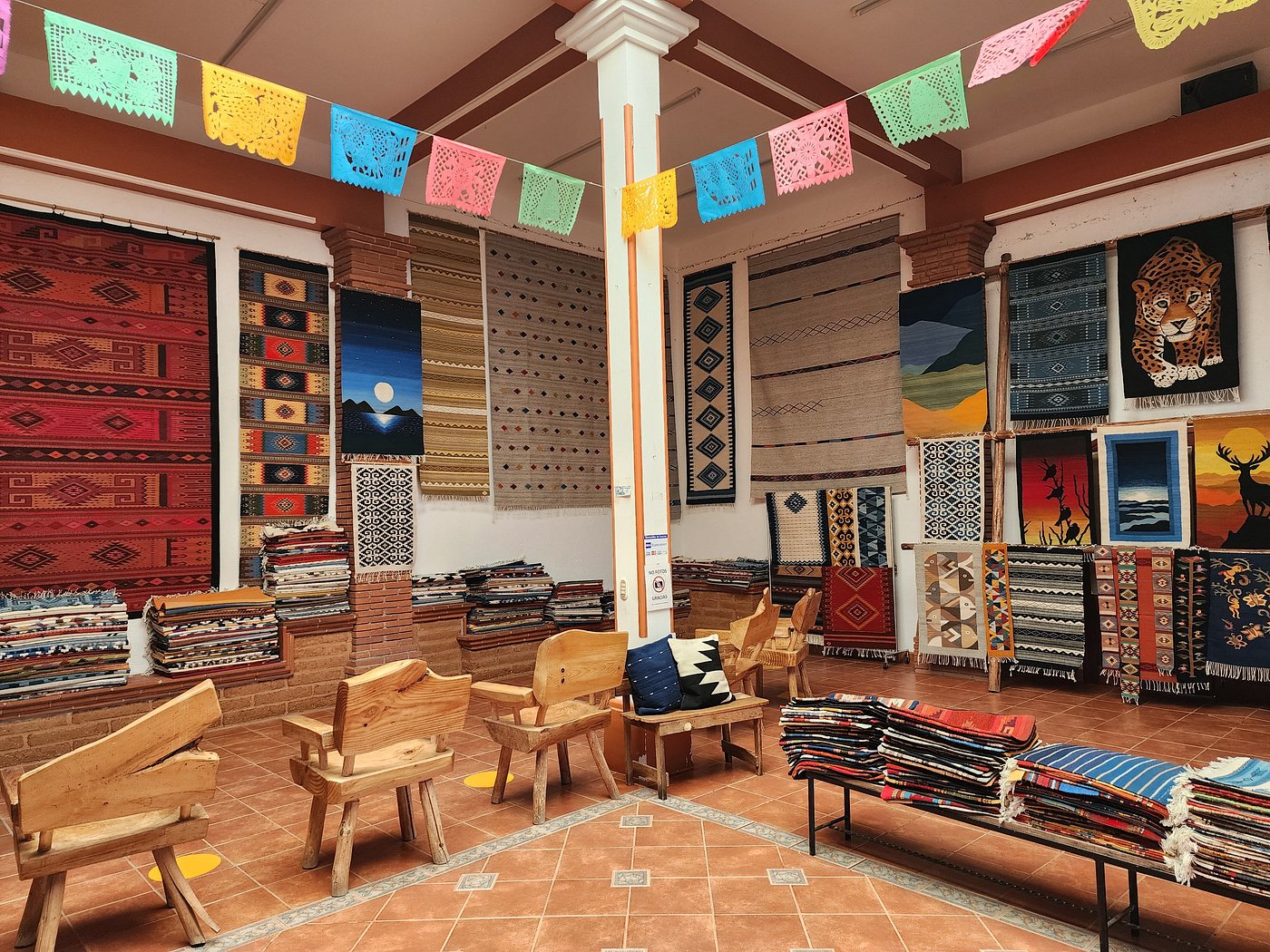 Papel picado overhead, finished tapetes floor to ceiling — every rug woven on-site in Teotitlán del Valle.